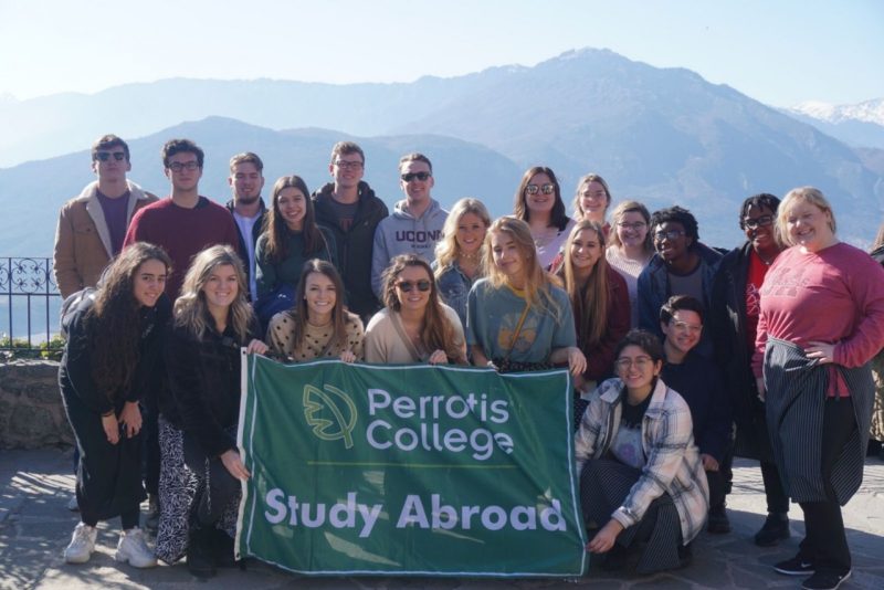 Students with mountains in the distance holding a banner that reads "Perrotis College Study Abroad".