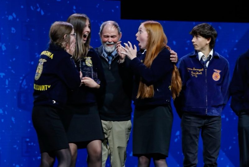 A group of girls reacting shock to winning an award on stage, with their FFA advisor.
