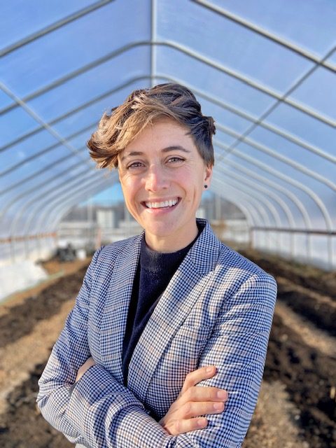 white woman with short hair in blue suite smiling, arms crossed in front of her chest, standing in a greenhouse.