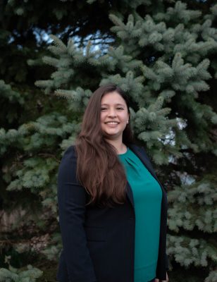 Woman with tan skin and long brown hair standing in front of trees. Wearing a green shirt and black blazer.