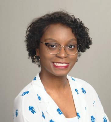 Black woman with short black hair and glasses, wearing a white blose with blue flowers on it