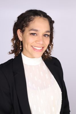 Woman with tan shin and black curly hair smiling, wearing a light top and black blazer