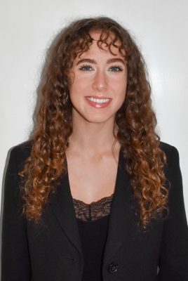 White woman with long curly brown/red hair smiling, wearing a black shirt and blazer