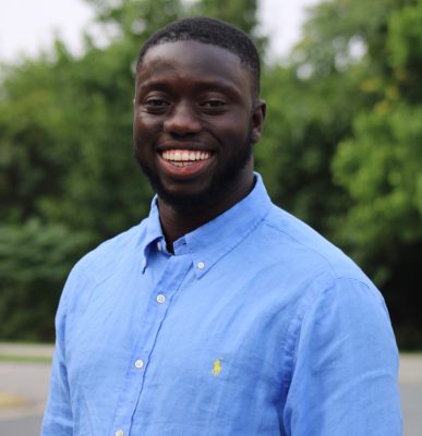 Black man smiling, standing outside wearing a blue button up shirt
