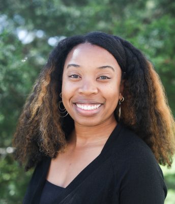 Black woman with natural hair standing outside in front of green leaves, smiling; wearing a black shirt. 