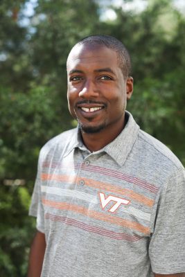 Black man wearing a grey polo shirt, standing outside smiling