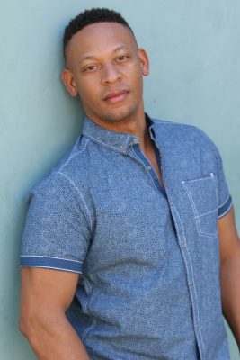 Black man leaning against a teal wall wearing a blue short sleeved button up shirt